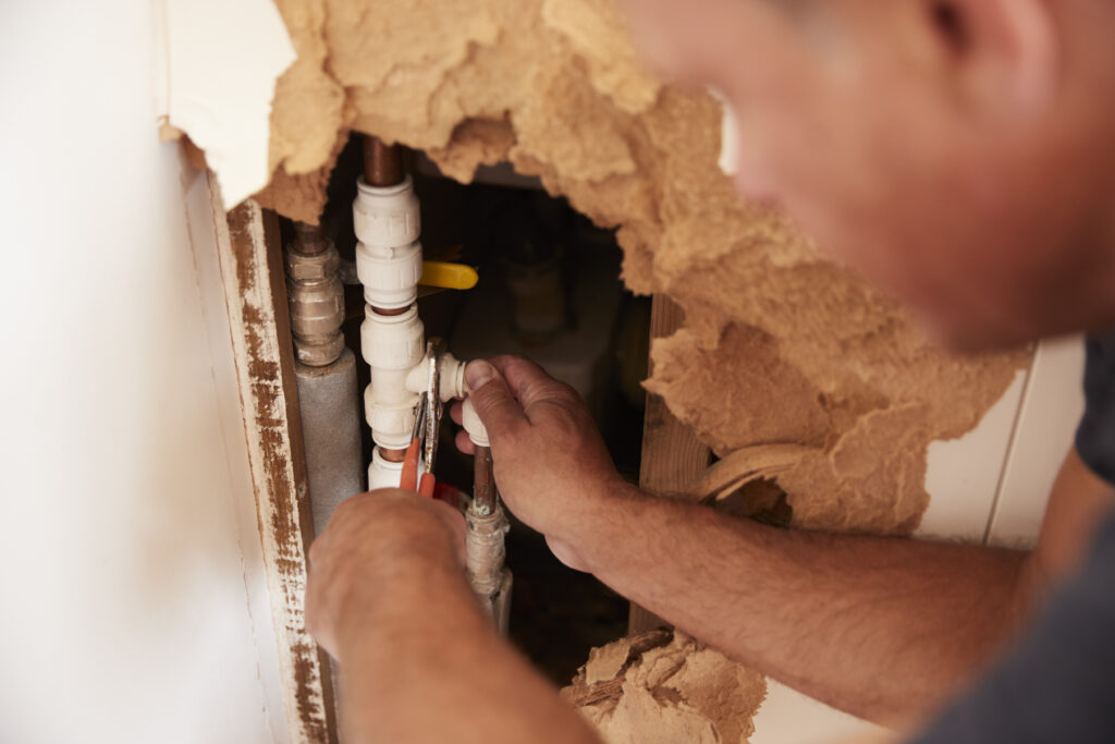 Over shoulder view of middle aged man repairing burst pipe and wall leak