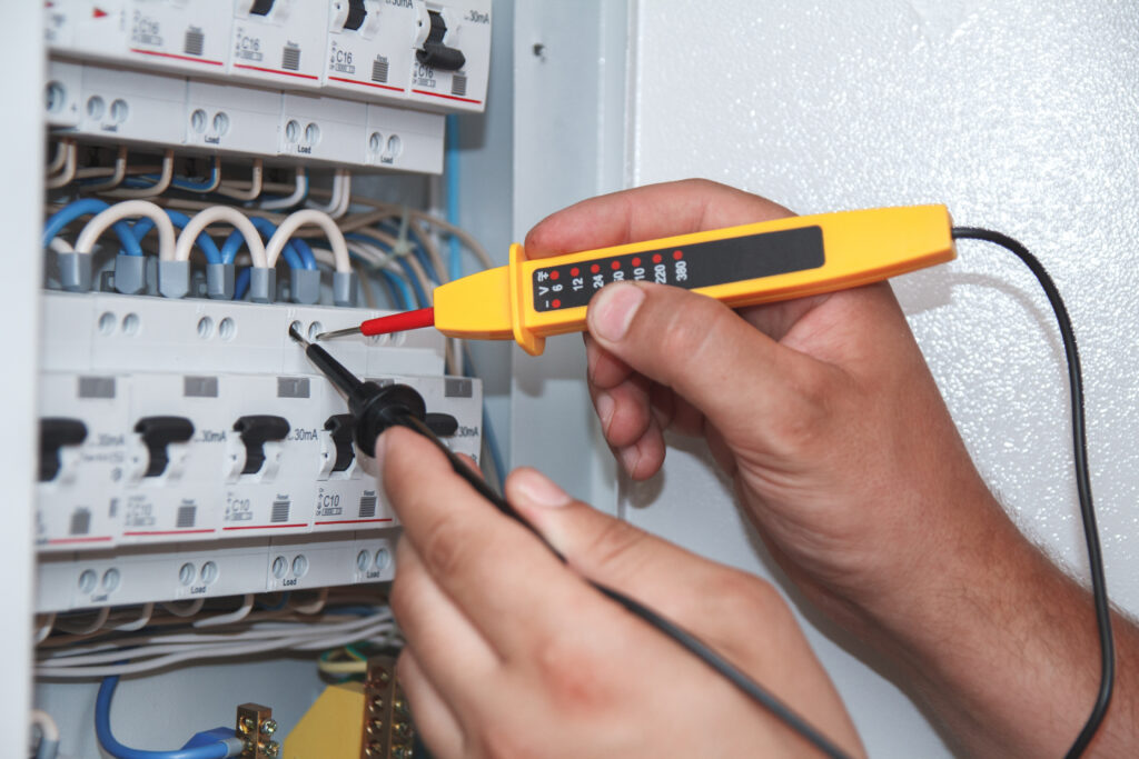 Hands of electrician with multimeter probe at an electrical switchgear cabinet examining fuse box during an electrical fault finding service